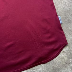 May include: A close-up shot of a maroon sports jersey. The fabric has a textured, breathable weave. A small tag with the word "HAMMERS" is visible on the lower right side. The jersey's hem is curved.