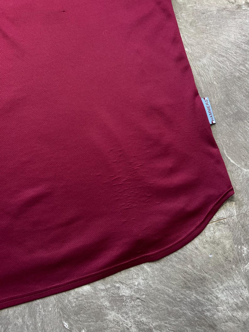 May include: A close-up shot of a maroon sports jersey. The fabric has a textured, breathable weave. A small tag with the word "HAMMERS" is visible on the lower right side. The jersey's hem is curved.