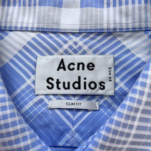 May include: A light blue and white plaid button-down shirt featuring a collar and a white fabric label stitched at the neck. The label displays the black text Acne Studios, Size 48, and a smaller tag below it reads SLIM FIT in black capital letters.