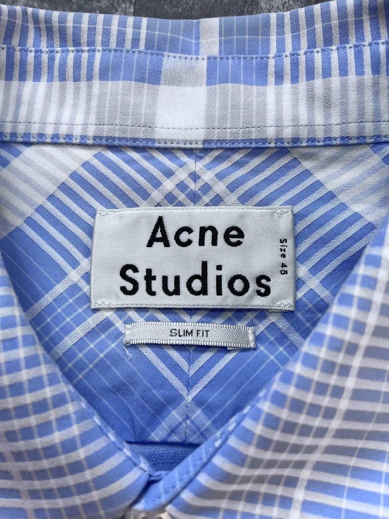 May include: A light blue and white plaid button-down shirt featuring a collar and a white fabric label stitched at the neck. The label displays the black text Acne Studios, Size 48, and a smaller tag below it reads SLIM FIT in black capital letters.