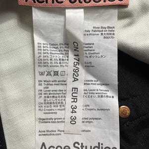 May include: A close-up of black jeans with a white interior. The label displays "Acne Studios" in pink, with size EUR 34 30. The jeans are made of cotton and polyester.