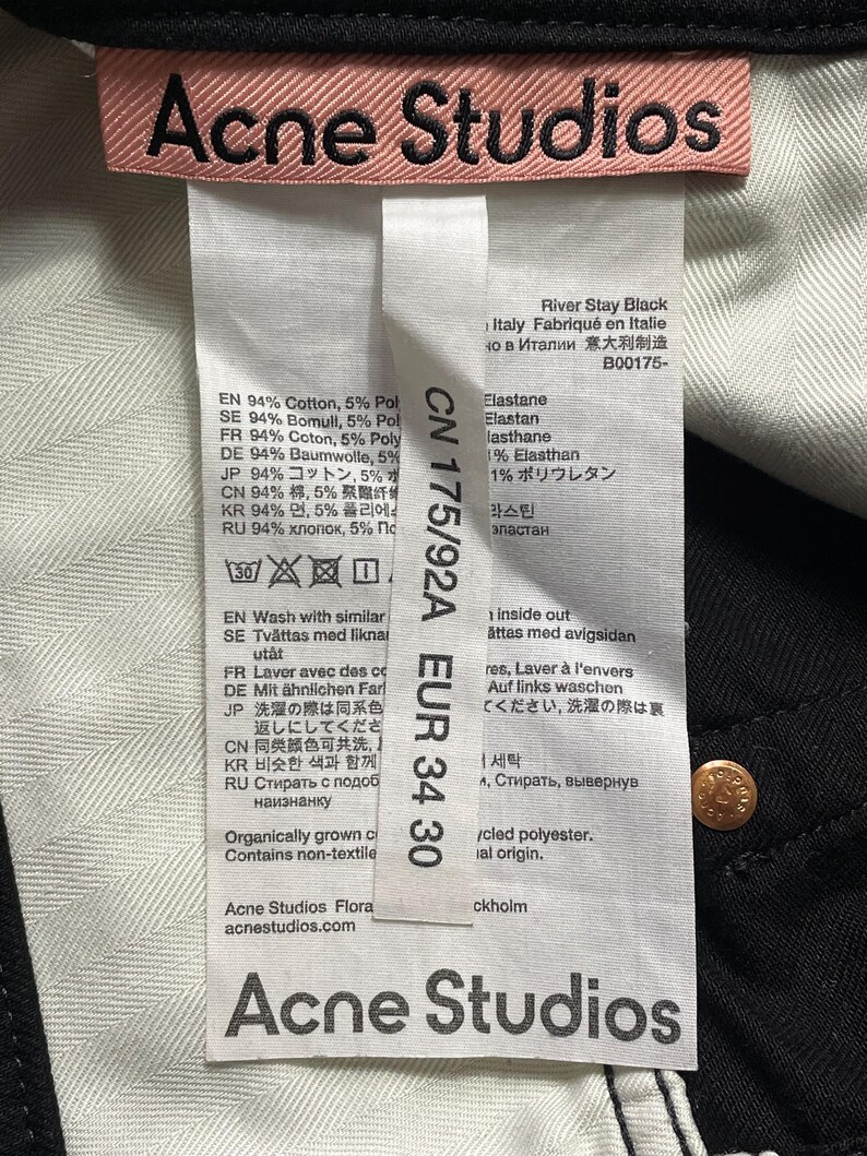 May include: A close-up of black jeans with a white interior. The label displays "Acne Studios" in pink, with size EUR 34 30. The jeans are made of cotton and polyester.