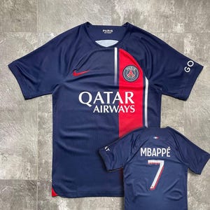 May include: A navy blue Paris Saint-Germain football shirt featuring a red vertical stripe, a white Qatar Airways logo, a Nike swoosh, and the PSG crest. A smaller view shows the back with the name MBAPP&Eacute; and the number 7 in white and red.