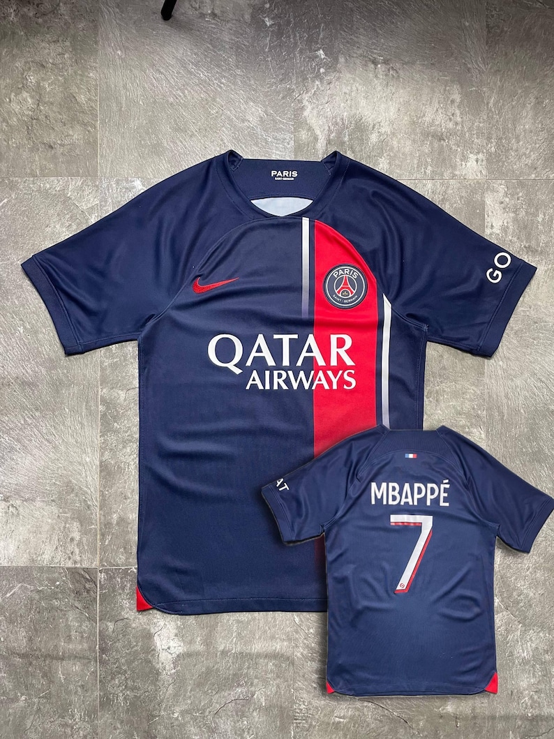 May include: A navy blue Paris Saint-Germain football shirt featuring a red vertical stripe, a white Qatar Airways logo, a Nike swoosh, and the PSG crest. A smaller view shows the back with the name MBAPP&Eacute; and the number 7 in white and red.
