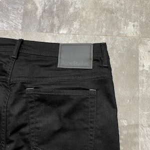 May include: Close-up of black denim jeans with a grey leather patch at the waistband. The patch features the brand name "Acne Studios". The jeans have a classic five-pocket design. The stitching is visible.