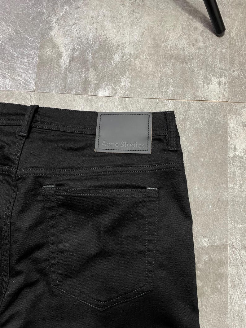 May include: Close-up of black denim jeans with a grey leather patch at the waistband. The patch features the brand name "Acne Studios". The jeans have a classic five-pocket design. The stitching is visible.
