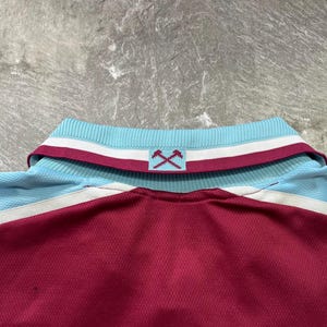 May include: Maroon and light blue polo shirt with a ribbed collar. The collar features white and maroon stripes, and a logo of crossed hammers. The shirt has a mesh-like texture.