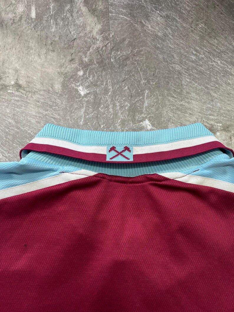 May include: Maroon and light blue polo shirt with a ribbed collar. The collar features white and maroon stripes, and a logo of crossed hammers. The shirt has a mesh-like texture.