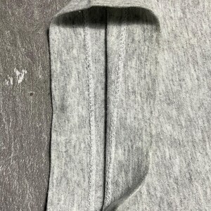 Puede incluir: Primer plano de una tela gris claro, posiblemente una camiseta u otra prenda. La imagen destaca la textura y los detalles de las costuras, con un enfoque en el borde doblado y las costuras. La tela parece suave y ligeramente texturizada.