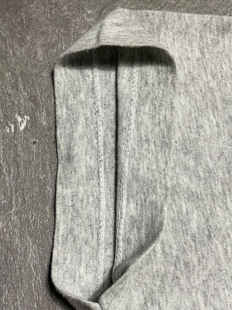 Puede incluir: Primer plano de una tela gris claro, posiblemente una camiseta u otra prenda. La imagen destaca la textura y los detalles de las costuras, con un enfoque en el borde doblado y las costuras. La tela parece suave y ligeramente texturizada.
