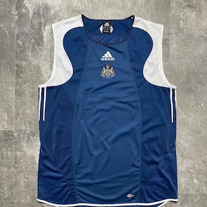 K&ouml;nnte beinhalten: Ein marineblaues Adidas &auml;rmelloses Trikot mit wei&szlig;en Akzenten. Das Trikot zeigt das Wappen von Newcastle United und das Adidas-Logo auf der Brust. Zwei wei&szlig;e Streifen verlaufen an den Seiten. Das Trikot hat einen runden Ausschnitt und eine meshartige Textur.