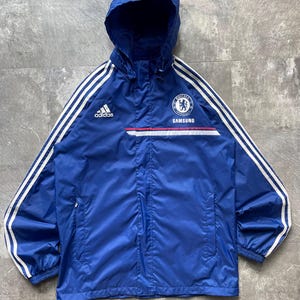 May include: A blue Adidas Chelsea FC hooded jacket with white stripes on the sleeves. The jacket features the Chelsea FC logo, the Adidas logo, and the Samsung logo. The jacket has a zipper closure and two front pockets.