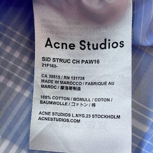May include: A white fabric care label attached to a light blue and white plaid shirt. The label features the text Acne Studios, SID STRUC CH PAW16, 21F163-, CA 39815 / RN 131738, MADE IN MAROCCO, 100% COTTON, and ACNE STUDIOS L.NYG.23 STOCKHOLM.
