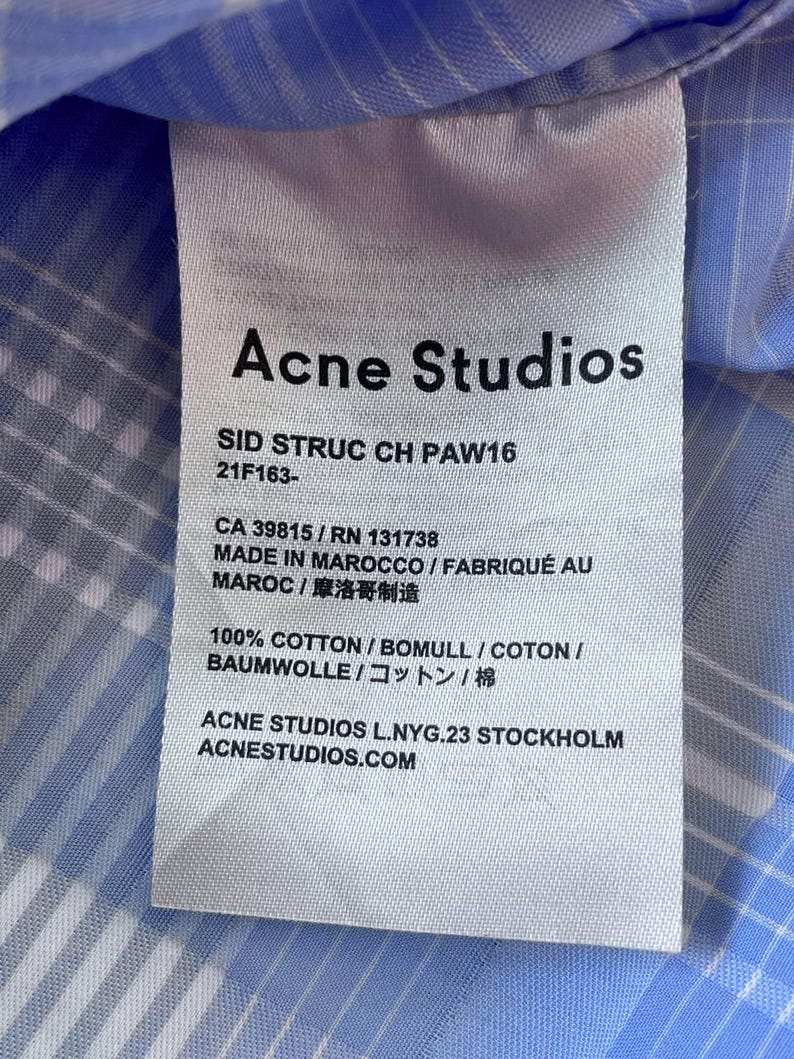 May include: A white fabric care label attached to a light blue and white plaid shirt. The label features the text Acne Studios, SID STRUC CH PAW16, 21F163-, CA 39815 / RN 131738, MADE IN MAROCCO, 100% COTTON, and ACNE STUDIOS L.NYG.23 STOCKHOLM.