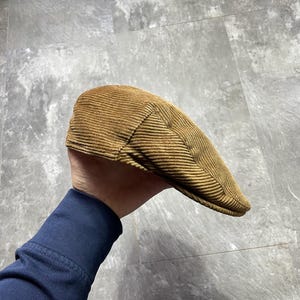 以下が含まれることがあります： 茶色のコーデュロイニュースボーイキャップ。キャップは暖かくアースカラーで、クラシックな丸みを帯びた形状とわずかに湾曲したつばが特徴です。コーデュロイ生地はリブ編みのテクスチャで、デザインに視覚的な面白さを加えています。スタイリッシュなアクセサリーです。