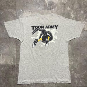 Puede incluir: Camiseta gris claro con el texto "TOON ARMY" sobre un dibujo animado. El dibujo animado presenta un personaje en blanco y negro con detalles amarillos, emergiendo de un dise&ntilde;o de nube. La camiseta est&aacute; tendida sobre una superficie gris.