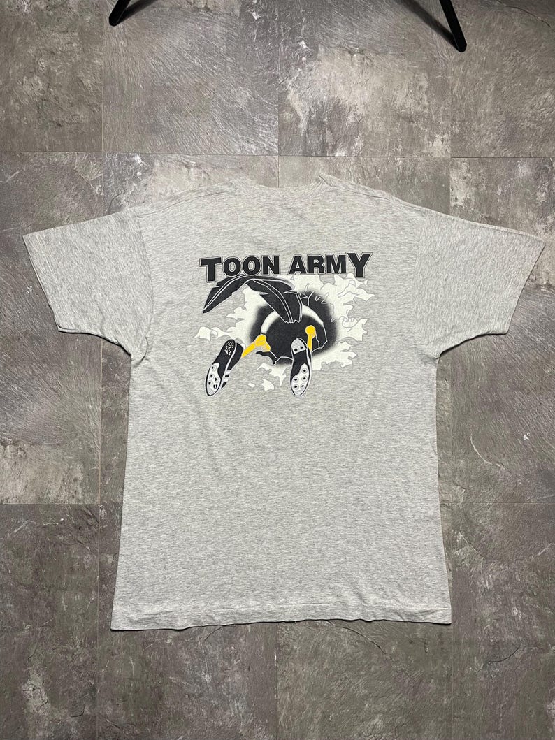 Puede incluir: Camiseta gris claro con el texto "TOON ARMY" sobre un dibujo animado. El dibujo animado presenta un personaje en blanco y negro con detalles amarillos, emergiendo de un dise&ntilde;o de nube. La camiseta est&aacute; tendida sobre una superficie gris.