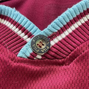 May include: Close-up of a maroon athletic jersey with a V-neck collar. The collar features light blue, white, and maroon stripes. A button with the brand name "FILA" is visible at the neckline. The jersey has a textured, breathable fabric.