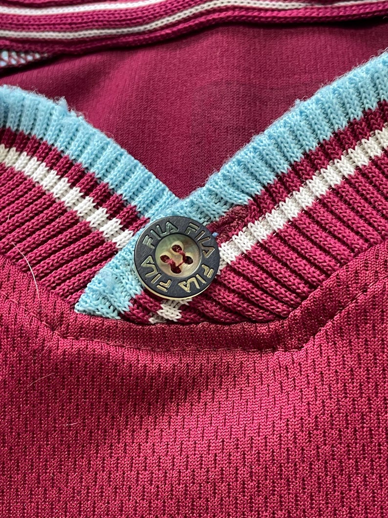 May include: Close-up of a maroon athletic jersey with a V-neck collar. The collar features light blue, white, and maroon stripes. A button with the brand name "FILA" is visible at the neckline. The jersey has a textured, breathable fabric.