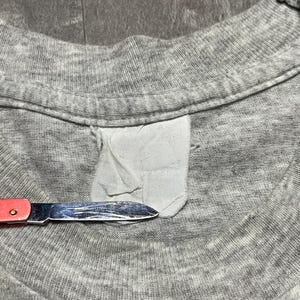 Puede incluir: Primer plano de un cuello de camiseta gris con una etiqueta blanca y un peque&ntilde;o cuchillo con mango rojo. El cuchillo est&aacute; cerca de la etiqueta, lo que sugiere una modificaci&oacute;n o reparaci&oacute;n.