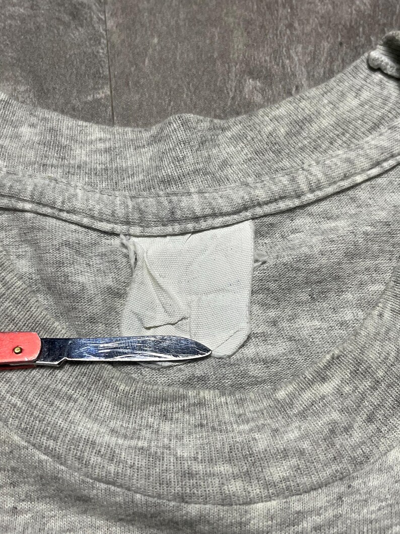 Puede incluir: Primer plano de un cuello de camiseta gris con una etiqueta blanca y un peque&ntilde;o cuchillo con mango rojo. El cuchillo est&aacute; cerca de la etiqueta, lo que sugiere una modificaci&oacute;n o reparaci&oacute;n.