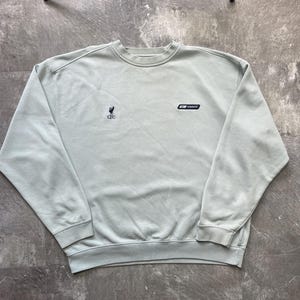 May include: A light sage green crewneck sweatshirt featuring a small dark navy blue Liverpool Football Club Liver bird logo on the left chest and a small dark navy blue Reebok brand logo patch on the right chest with long sleeves and ribbed cuffs.