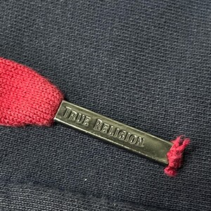May include: Close-up of a navy blue fabric with a red drawstring. A silver metal tag with the words "TRUE RELIGION" is attached to the drawstring. The fabric has a textured appearance.