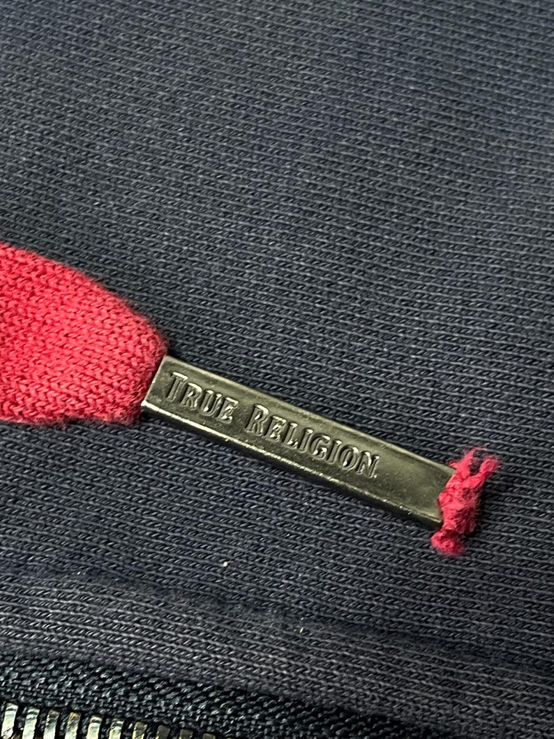 May include: Close-up of a navy blue fabric with a red drawstring. A silver metal tag with the words "TRUE RELIGION" is attached to the drawstring. The fabric has a textured appearance.
