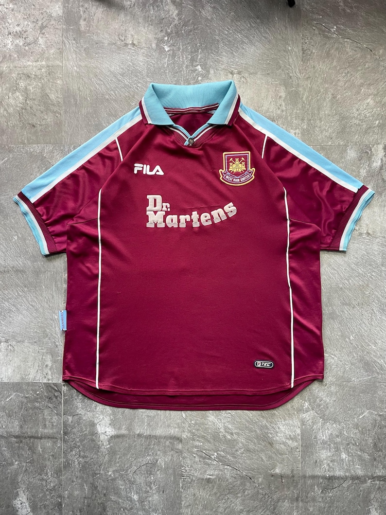 May include: Maroon football shirt with light blue collar and sleeve trim. The shirt displays the Fila logo, the Dr. Martens logo, and a team crest. White stripes run down the sides of the shirt.
