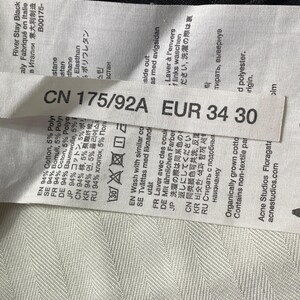 May include: A close-up of a label on black denim jeans. The label shows the size EUR 34 30, and fabric details. The jeans have a gold button. The label also includes the text "River Stay Black".