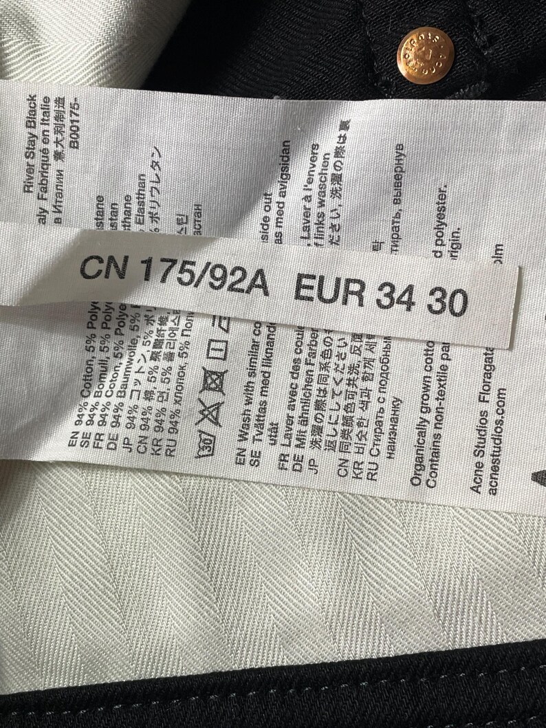 May include: A close-up of a label on black denim jeans. The label shows the size EUR 34 30, and fabric details. The jeans have a gold button. The label also includes the text "River Stay Black".