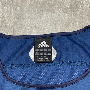 K&ouml;nnte beinhalten: Ein marineblaues Adidas-Sporttrikot mit wei&szlig;em Adidas-Logo und einem Etikett mit der Aufschrift "Made in China". Das Trikot hat eine Mesh-Struktur und eine dunkelblaue Einfassung.