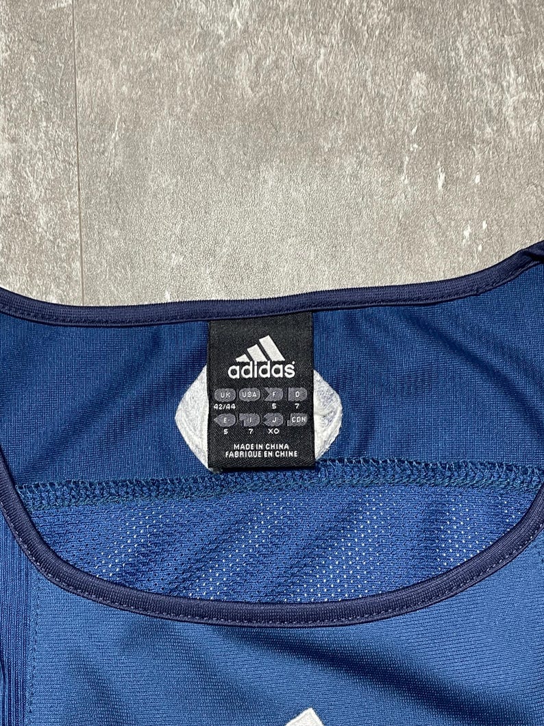 K&ouml;nnte beinhalten: Ein marineblaues Adidas-Sporttrikot mit wei&szlig;em Adidas-Logo und einem Etikett mit der Aufschrift "Made in China". Das Trikot hat eine Mesh-Struktur und eine dunkelblaue Einfassung.