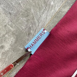 May include: A close-up shot of maroon fabric with a white stripe and a light blue tag with the word "HAMMERS" in white. A pair of scissors with a red handle is cutting the tag.