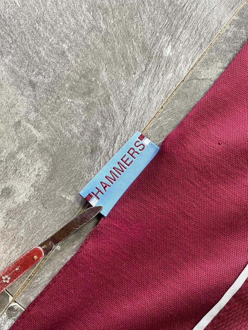 May include: A close-up shot of maroon fabric with a white stripe and a light blue tag with the word "HAMMERS" in white. A pair of scissors with a red handle is cutting the tag.