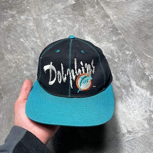 Puede incluir: Gorra de béisbol negra y turquesa de los Dolphins. La gorra presenta el nombre del equipo en escritura blanca y un logotipo de un delfín. La visera es turquesa y la parte superior de la gorra es negra.