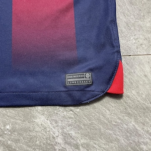 May include: A navy blue athletic jersey featuring a vertical maroon stripe and a small red accent at the hem. A black rectangular tag is stitched near the bottom corner with the text ENGINEERED and the alphanumeric code 128623AAC2 printed on it.