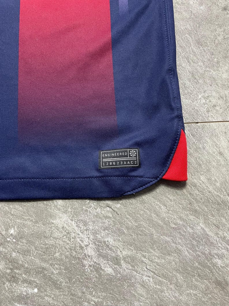 May include: A navy blue athletic jersey featuring a vertical maroon stripe and a small red accent at the hem. A black rectangular tag is stitched near the bottom corner with the text ENGINEERED and the alphanumeric code 128623AAC2 printed on it.