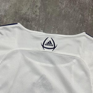 Puede incluir: Camiseta deportiva blanca con el logo de Adidas en azul marino. La camiseta tiene un panel de malla en la espalda y tejido acanalado en los hombros. El cuello est&aacute; ribeteado en azul marino.