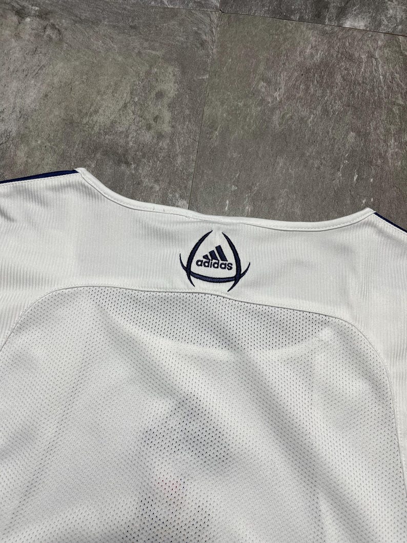 Puede incluir: Camiseta deportiva blanca con el logo de Adidas en azul marino. La camiseta tiene un panel de malla en la espalda y tejido acanalado en los hombros. El cuello est&aacute; ribeteado en azul marino.