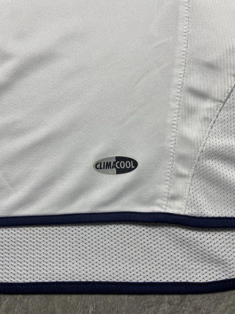 Puede incluir: Camiseta deportiva blanca con ribete azul marino y el logo "CLIMACOOL". La camiseta tiene un panel de malla en el lateral y una secci&oacute;n inferior perforada. La tela parece ser un material ligero y transpirable.