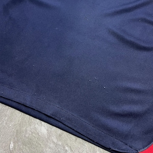 May include: A dark blue athletic jersey featuring a red and white geometric accent at the top and a small red trim at the bottom corner. The fabric has a smooth texture with visible signs of minor wear, including small snags on the surface.