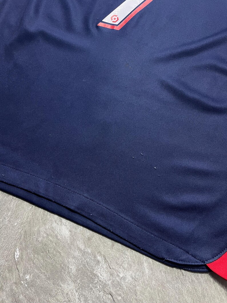 May include: A dark blue athletic jersey featuring a red and white geometric accent at the top and a small red trim at the bottom corner. The fabric has a smooth texture with visible signs of minor wear, including small snags on the surface.