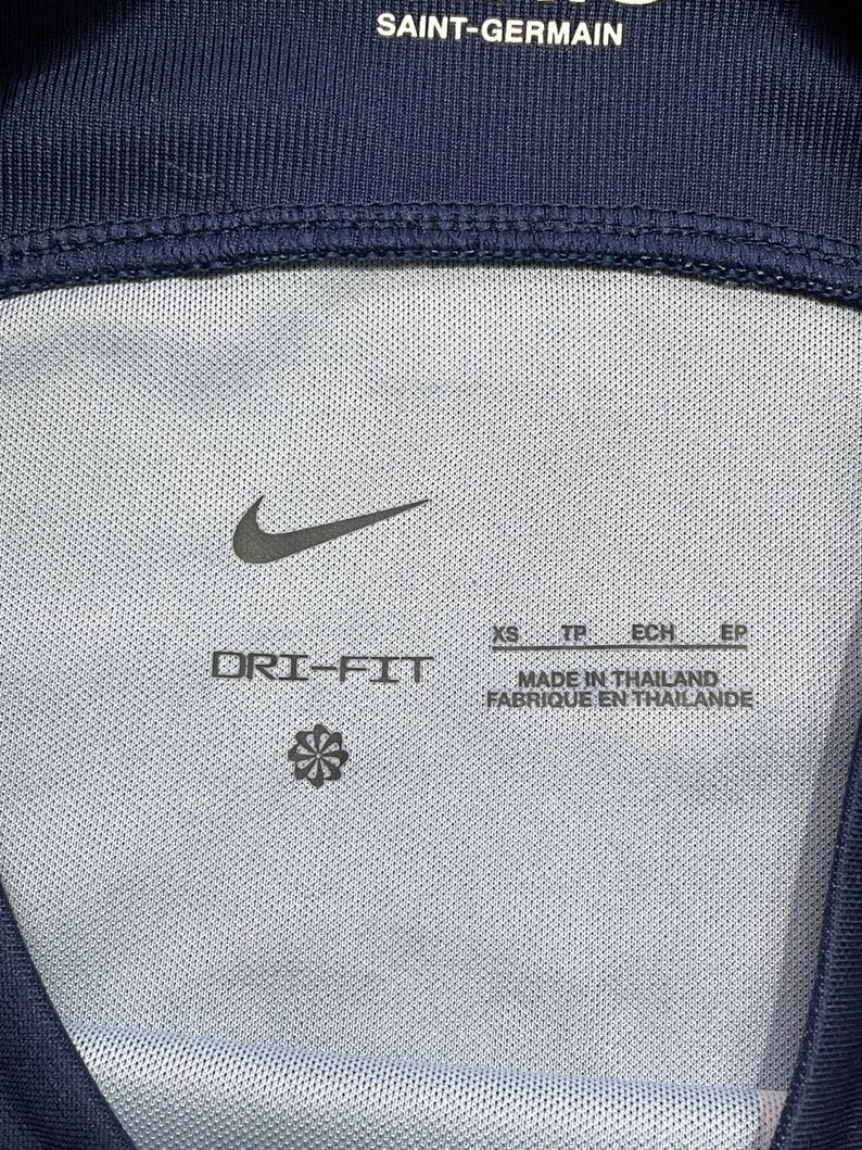 May include: A navy blue athletic garment interior featuring the text SAINT-GERMAIN, a black Nike swoosh logo, and the text DRI-FIT. The label indicates size XS, TP, ECH, EP and states MADE IN THAILAND, FABRIQUE EN THAILANDE with a small flower icon.