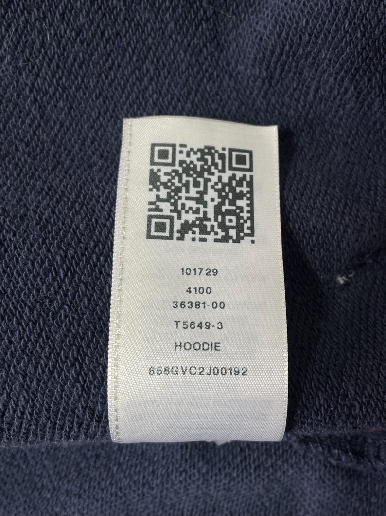 May include: Close-up of a navy blue fabric with a white tag attached. The tag features a QR code and printed text: 101729, 4100, 36381-00, T5649-3, HOODIE, and 858GVC2J00192. The fabric has a textured appearance.