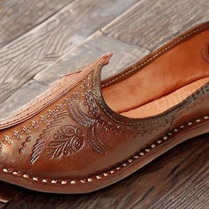 May include: A brown leather slip-on shoe featuring an intricate embossed floral pattern on the upper, a pointed toe, and a visible white stitched border along the sole. The interior lining is tan, and a logo is printed on the heel area.