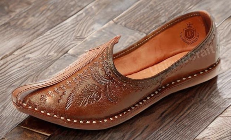 May include: A brown leather slip-on shoe featuring an intricate embossed floral pattern on the upper, a pointed toe, and a visible white stitched border along the sole. The interior lining is tan, and a logo is printed on the heel area.