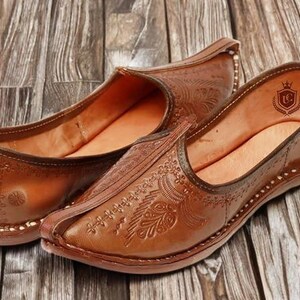 May include: A pair of tan leather slip-on shoes featuring an upturned pointed toe and intricate embossed floral patterns across the upper. The interior lining is a lighter tan shade and displays a small shield-shaped logo with a crown emblem.