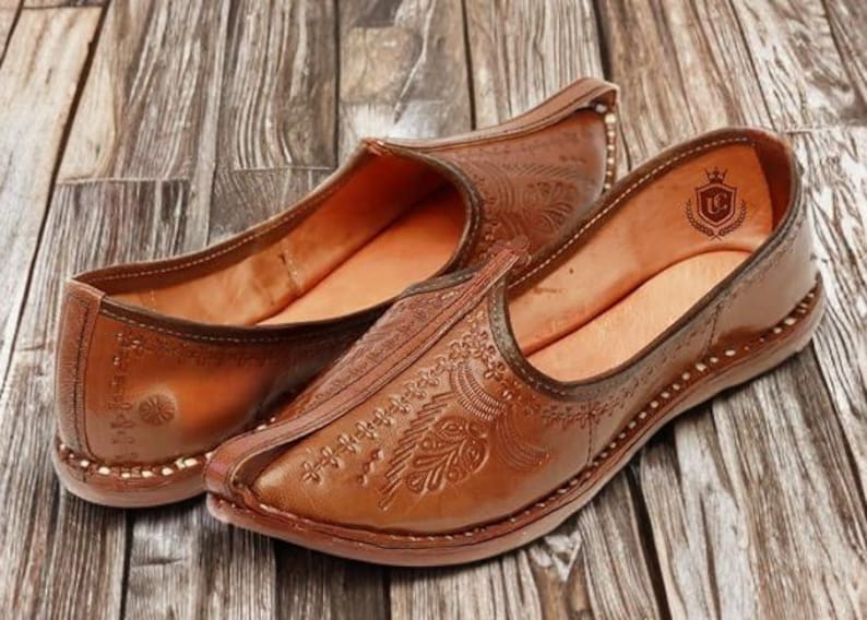 May include: A pair of tan leather slip-on shoes featuring an upturned pointed toe and intricate embossed floral patterns across the upper. The interior lining is a lighter tan shade and displays a small shield-shaped logo with a crown emblem.