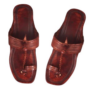 May include: A pair of dark brown leather sandals featuring a wide strap across the top with intricate embossed detailing, a central braided leather cord, and a toe loop. The footbed has a stitched border and a smooth, polished finish throughout.